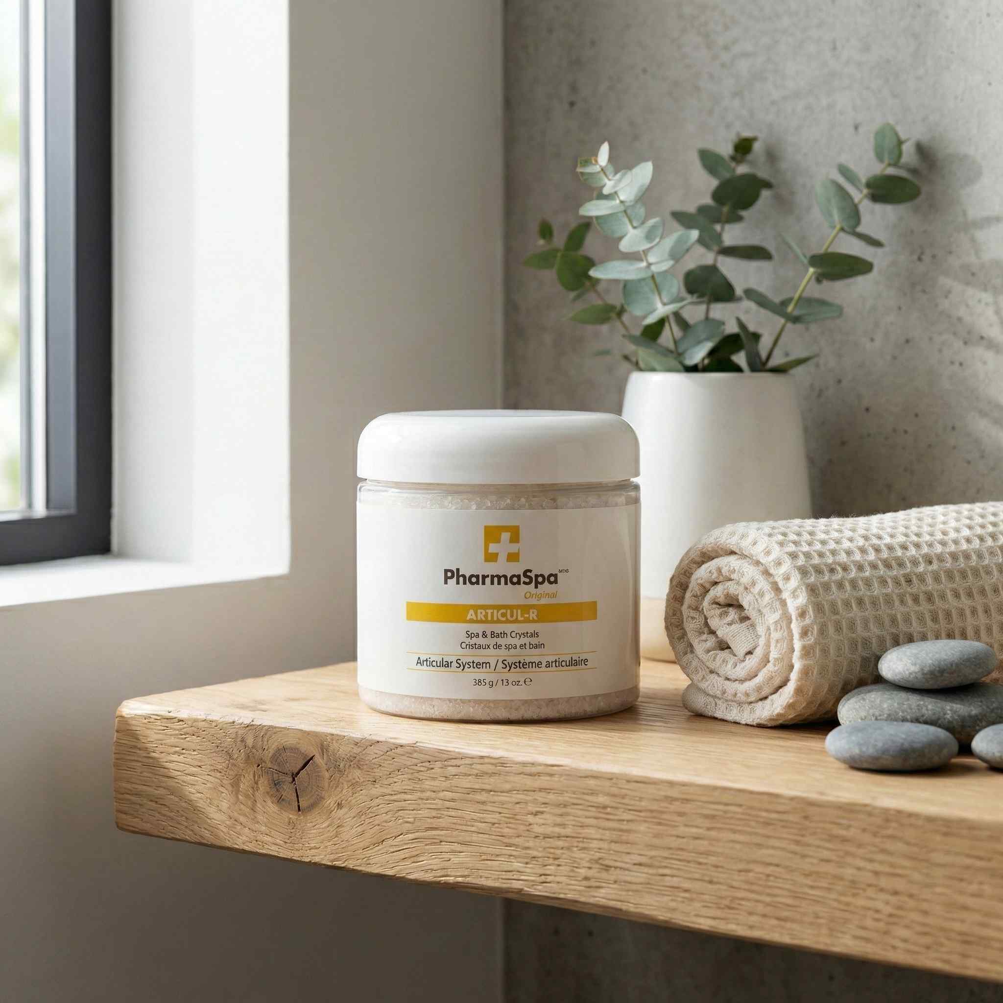 White jar of PharmaSpa Articul-R spa crystals displayed with eucalyptus sprigs, a rolled towel, and spa accessories