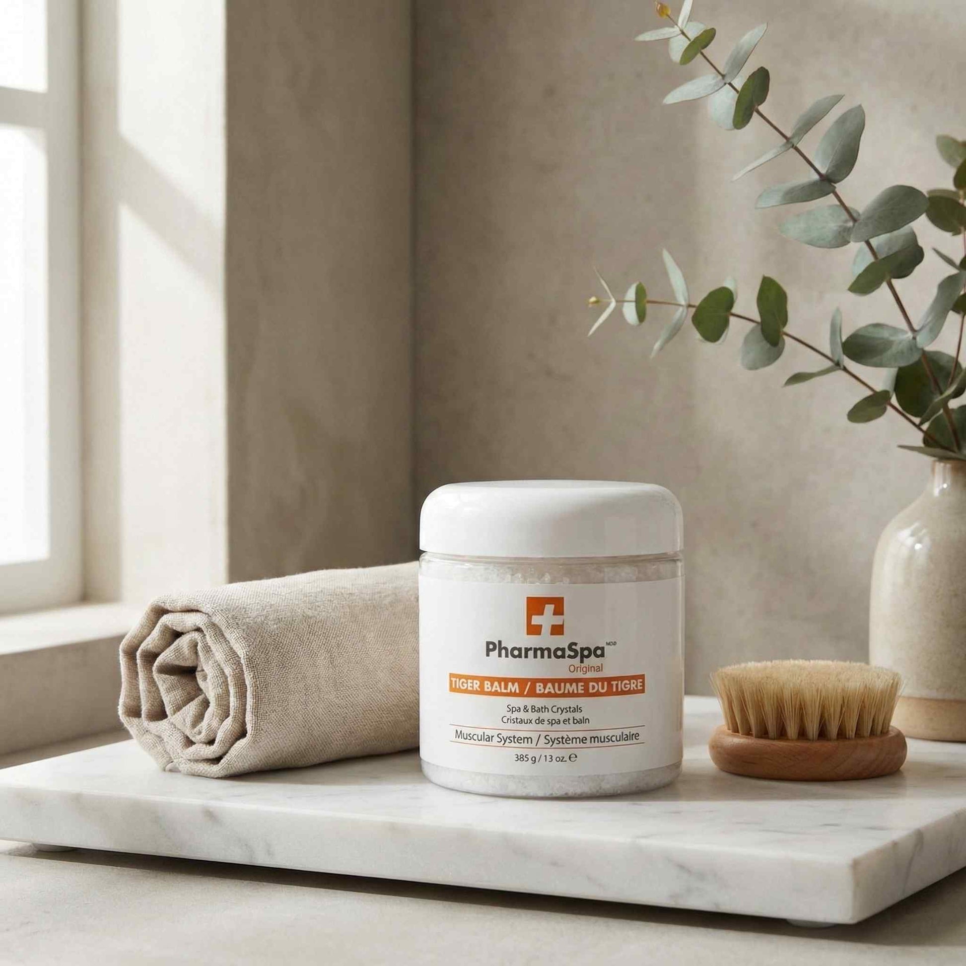 White jar of PharmaSpa Tiger Balm spa and bath crystals on a marble surface with a towel and wooden brush in a spa setting