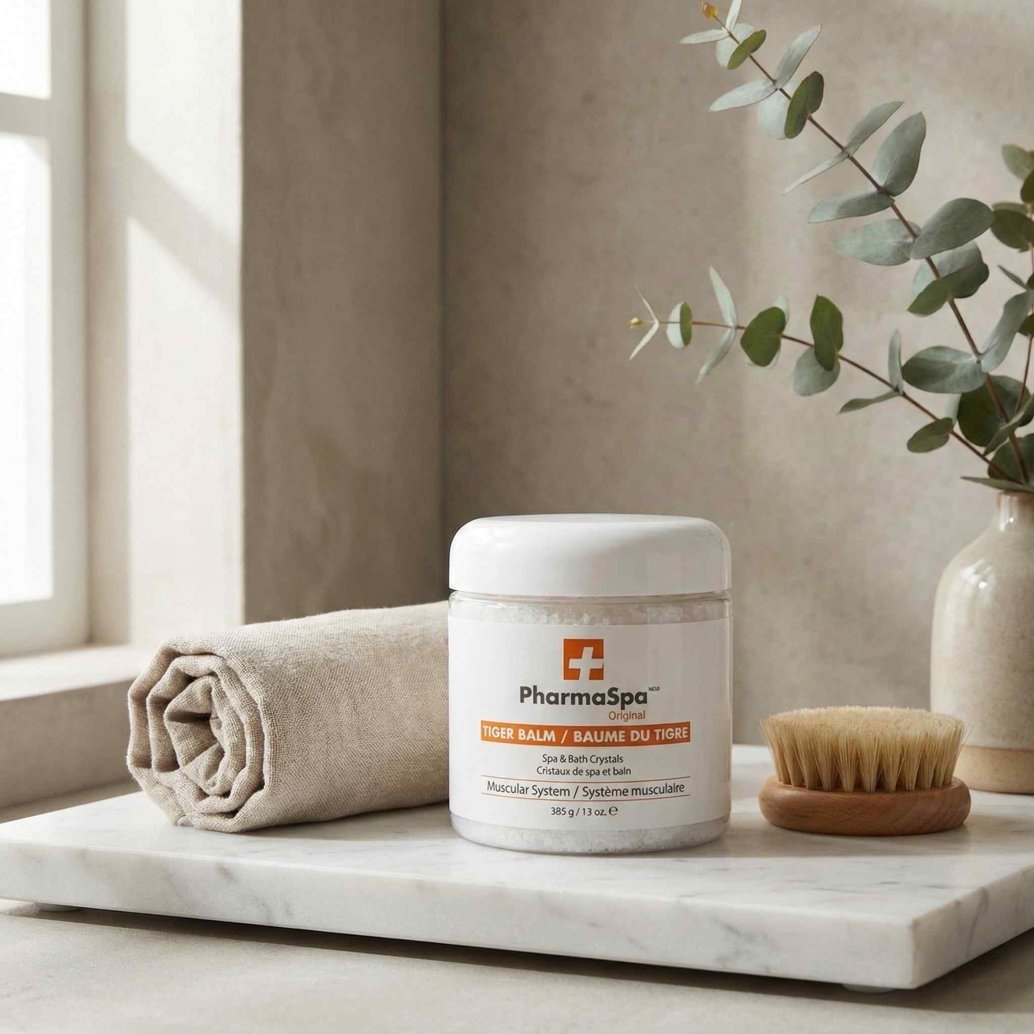 White jar of PharmaSpa Tiger Balm spa and bath crystals on a marble surface with a towel and wooden brush in a spa setting